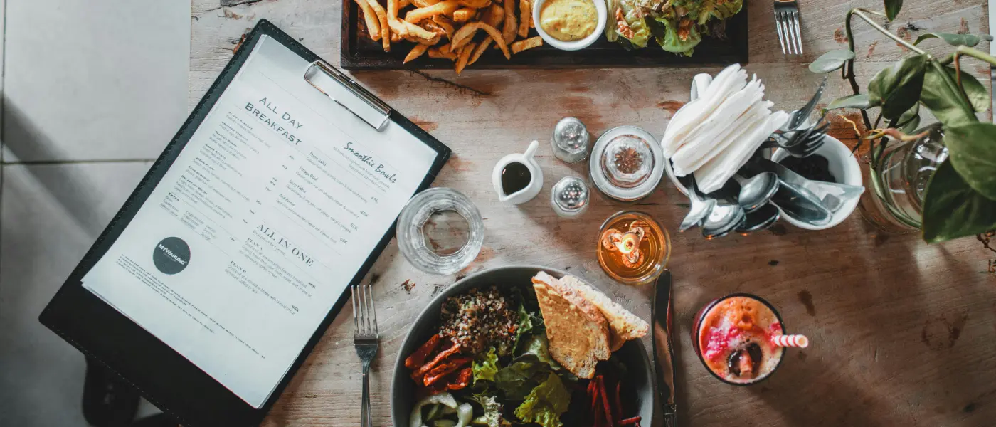 How to Design the Perfect Restaurant Menu - a variety of food and a stark white menu on a clipboard are seen on a table from a bird's eye view.